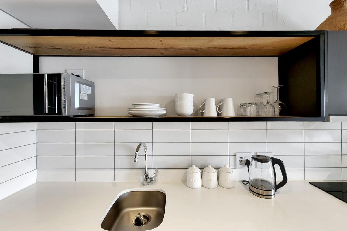 Modern kitchen area featuring a clean subway tile backsplash, sleek quartz countertops, an undermount sink, and stylish open shelving against an exposed brick wall.