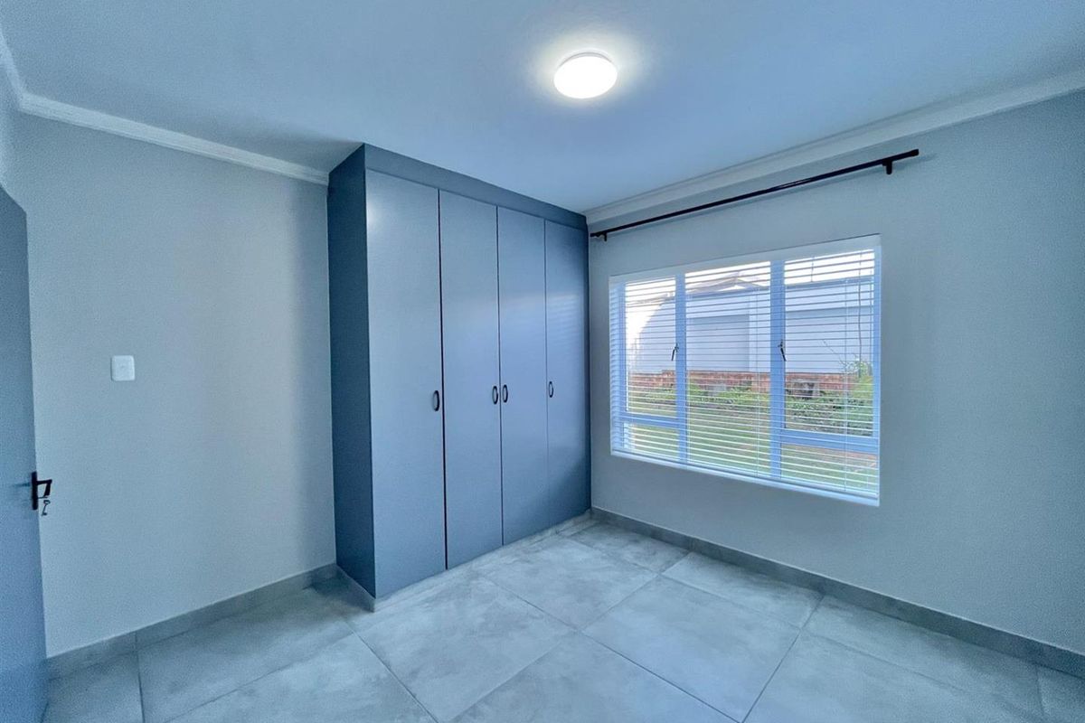 A bright, empty bedroom featuring modern grey floor tiles, built-in cupboards, and a large window with fitted blinds.