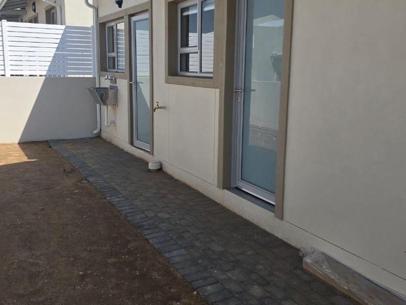 A rear exterior view showing a paved walkway, external sink, and white privacy fencing, with an unfinished garden area.