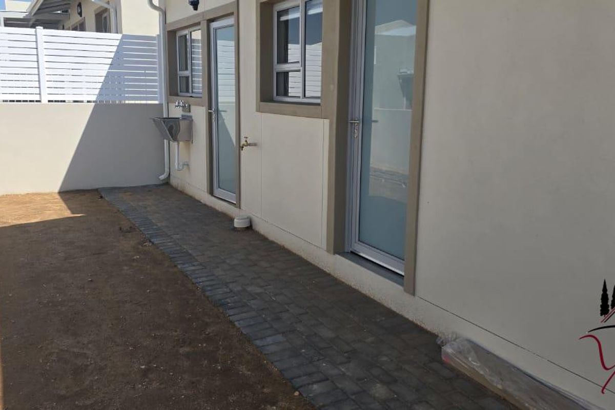 A rear exterior view showing a paved walkway, external sink, and white privacy fencing, with an unfinished garden area.