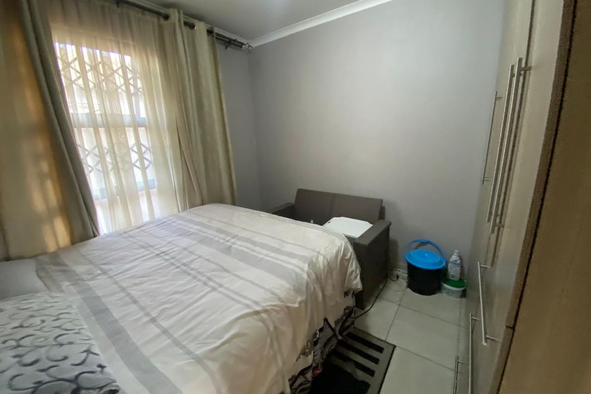 A compact bedroom featuring tiled flooring, built-in cupboards, and a window fitted with security bars and curtains.