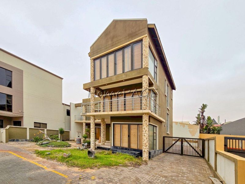 Three-story modern residential property featuring stone-clad columns, metal balcony railings, and a paved driveway with secure gated access.