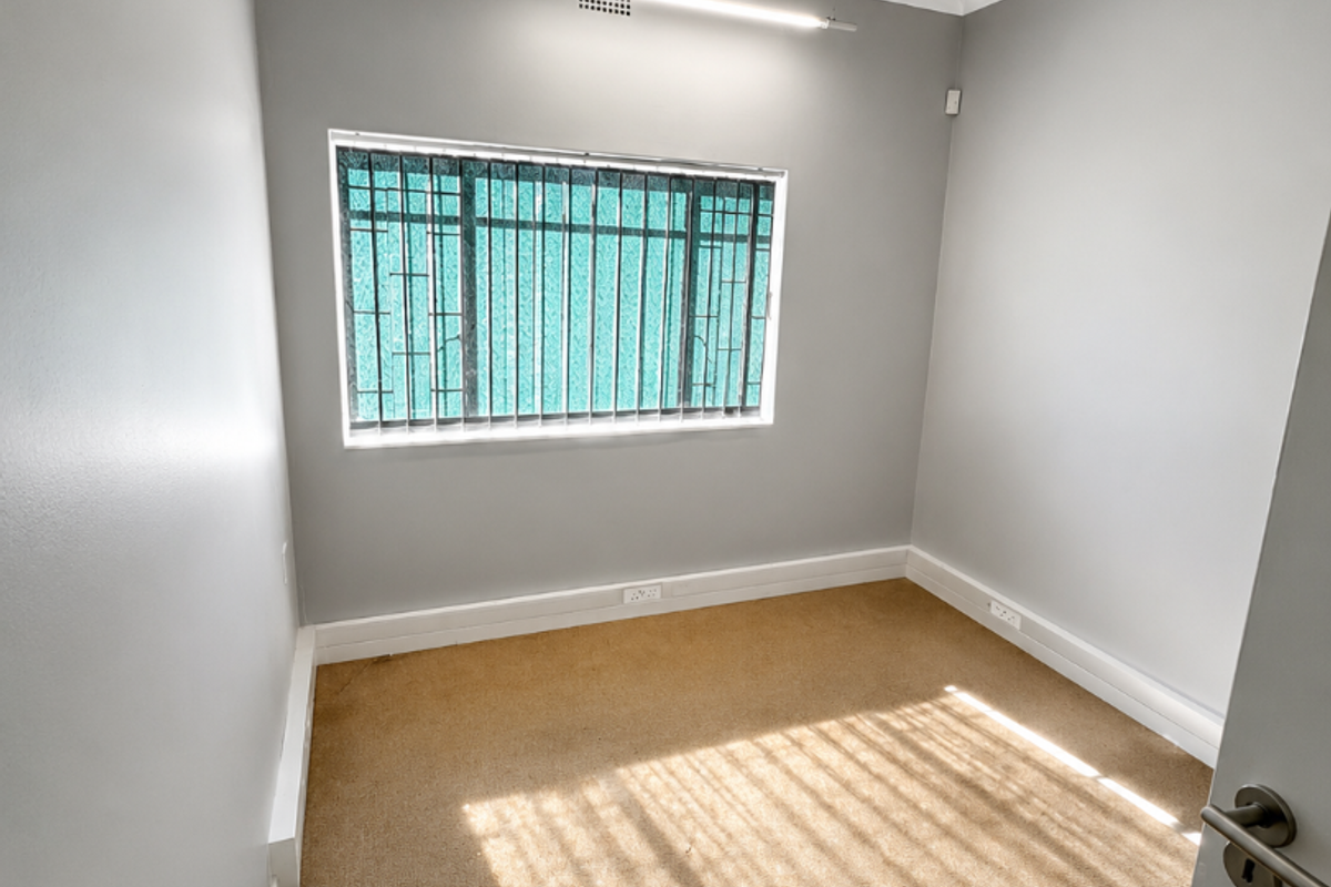 A bright, empty bedroom featuring neutral grey walls, carpeted flooring, and a window fitted with security bars.