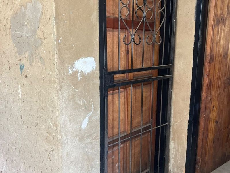 Exterior entrance area featuring a wooden door with a black metal security gate, showing signs of wear on the surrounding concrete wall.