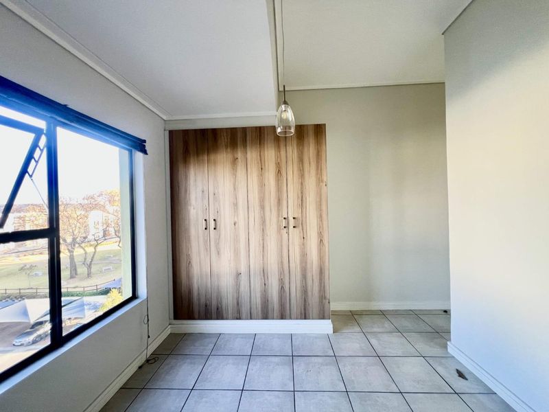 A bright, empty bedroom featuring modern wood-finish built-in cupboards, neutral tiled flooring, and a large window providing natural light.