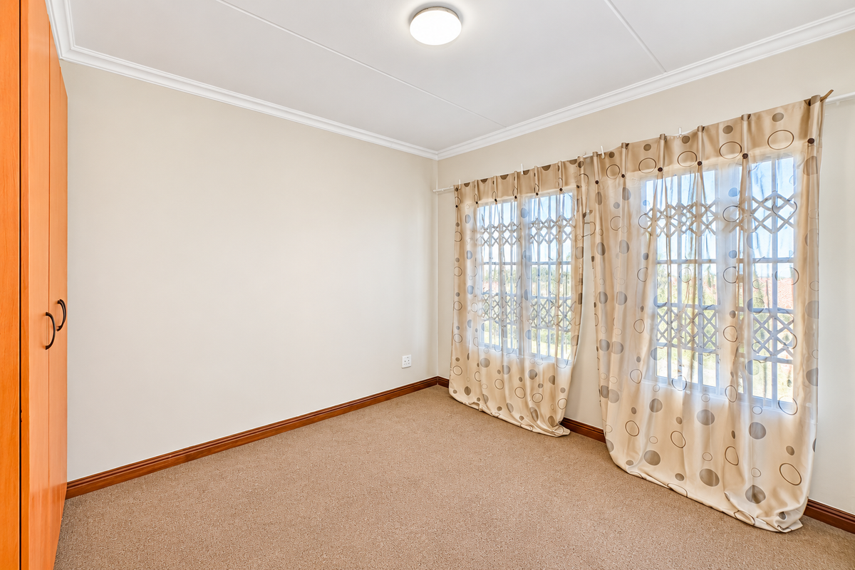 A bright, empty bedroom featuring neutral walls, carpeted flooring, a built-in wardrobe, and windows fitted with security gates.