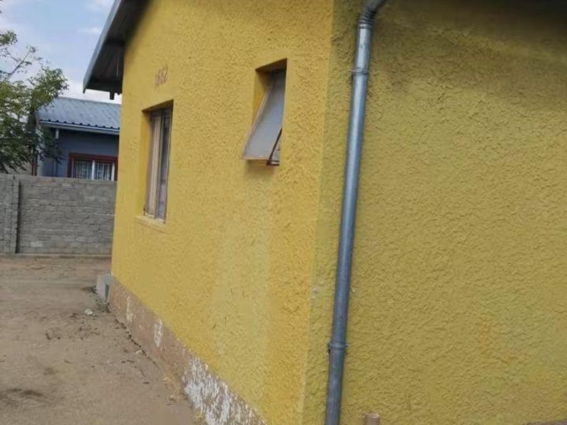 Side view of the property showing textured yellow plaster walls, metal roofing, and external plumbing infrastructure with visible signs of wear and damp.
