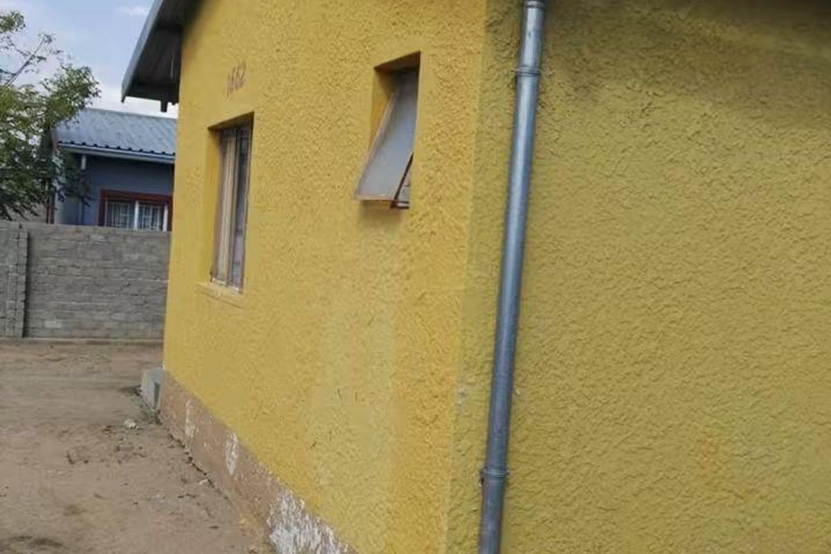 Side view of the property showing textured yellow plaster walls, metal roofing, and external plumbing infrastructure with visible signs of wear and damp.