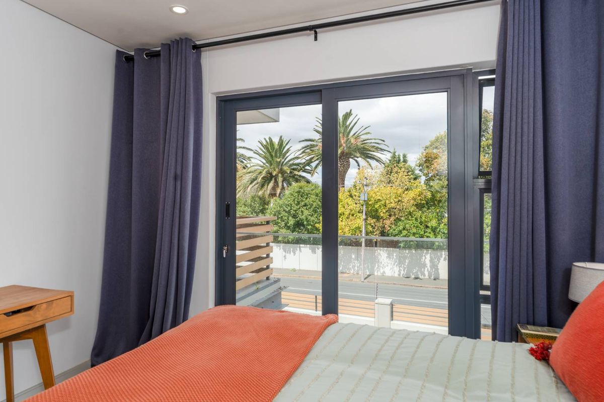 A bright bedroom featuring modern dark-framed sliding glass doors that open onto a private balcony with pleasant street and tree-lined views.