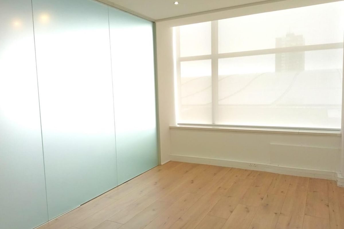 A bright, modern office space featuring timber laminate flooring, a sleek frosted glass partition, and large windows with integrated roller blinds.