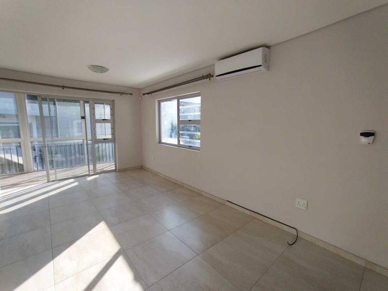 A bright, empty living room featuring neutral tiled flooring, a wall-mounted air conditioning unit, and large sliding glass doors leading to an outdoor area.