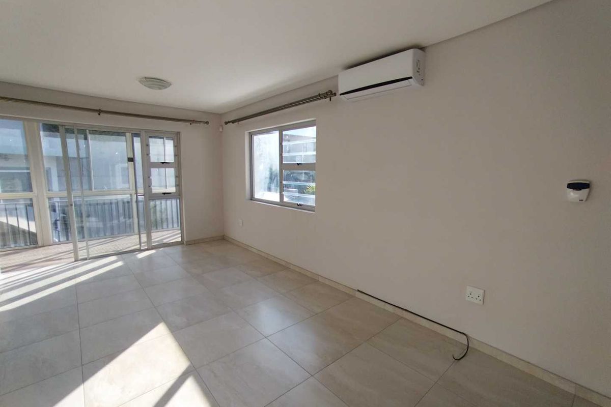 A bright, empty living room featuring neutral tiled flooring, a wall-mounted air conditioning unit, and large sliding glass doors leading to an outdoor area.