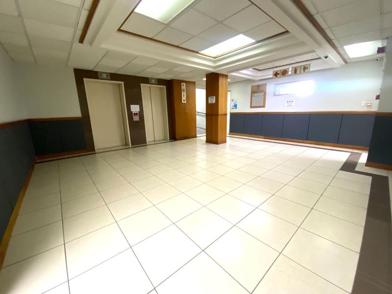 A spacious apartment building lobby or hallway featuring dual elevators, tiled flooring, and a suspended ceiling with recessed lighting.