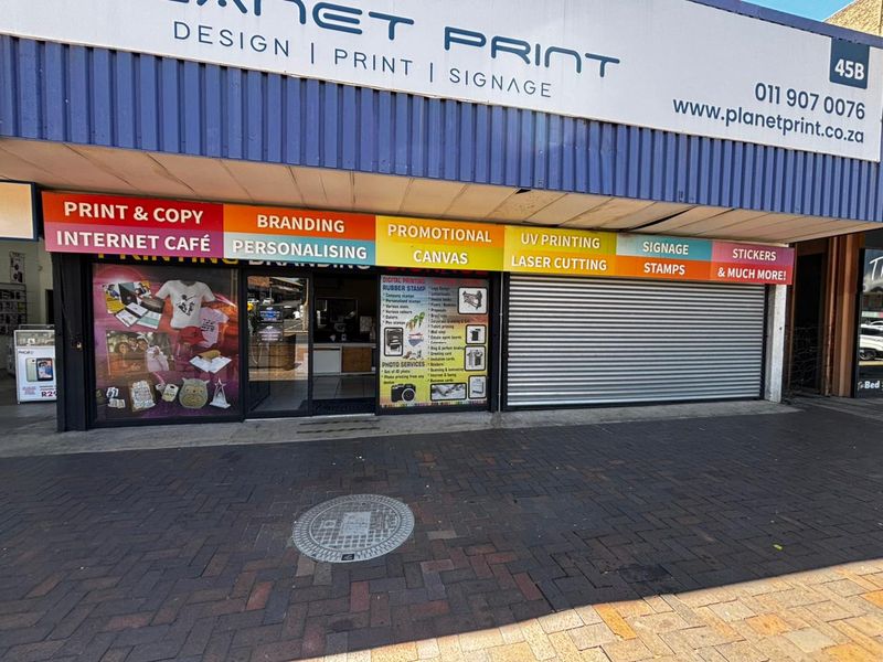 Front facade of a commercial print and signage shop featuring large storefront signage, glass entrance, and a secure metal roller shutter.