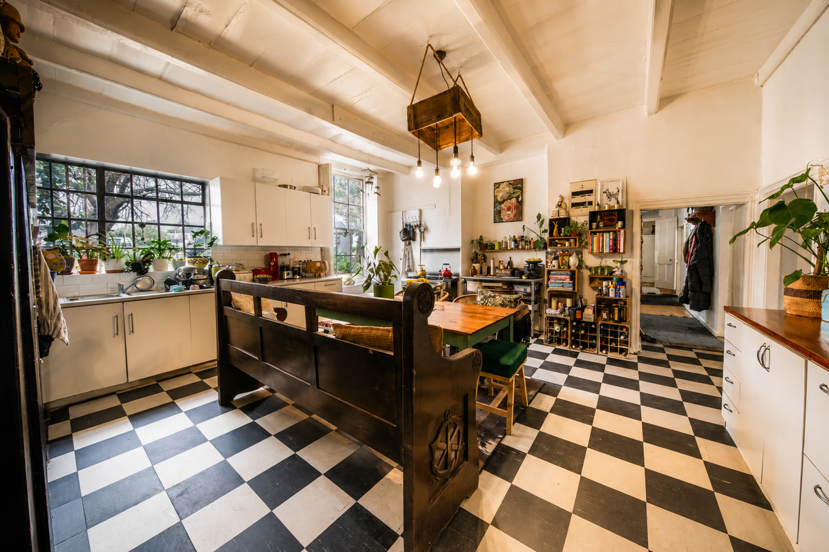 A rustic kitchen featuring exposed timber ceiling beams, checkerboard flooring, white cabinetry, and a tiled backsplash, offering a character-filled space with potential for modernization.