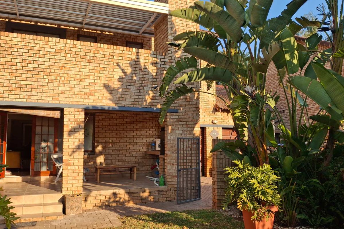 A rear view of the townhouse featuring a face brick facade, a covered patio area with a built-in braai, and a paved courtyard.