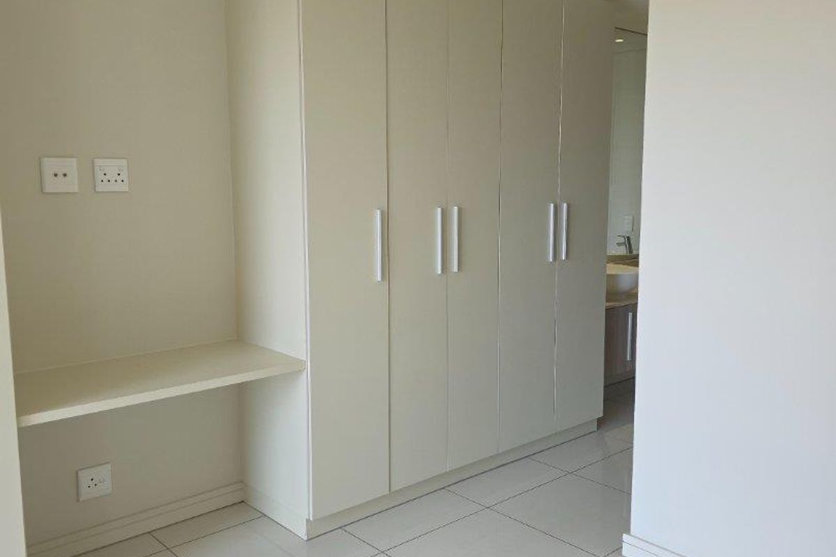 A bright bedroom featuring built-in floor-to-ceiling wardrobes and an integrated desk area, finished with neutral walls and clean tiled flooring.