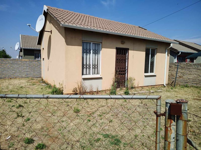 Front view of a detached single-story house featuring a tiled roof, security gates, and burglar bars, set within a fenced yard with overgrown vegetation.