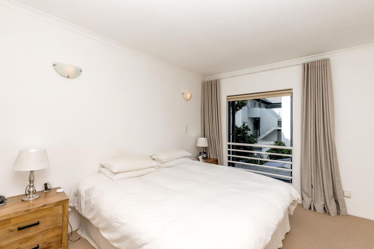 A well-maintained bedroom featuring neutral carpet flooring, wall-mounted lighting, and a window with a safety railing overlooking the exterior.