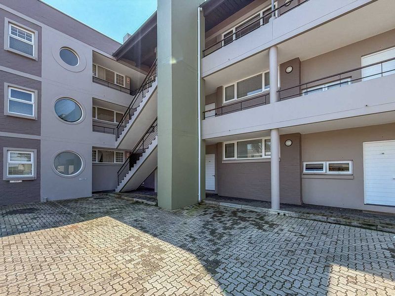 Exterior view of an apartment complex featuring a paved courtyard, external staircase, and multi-level balconies with circular and standard window designs.