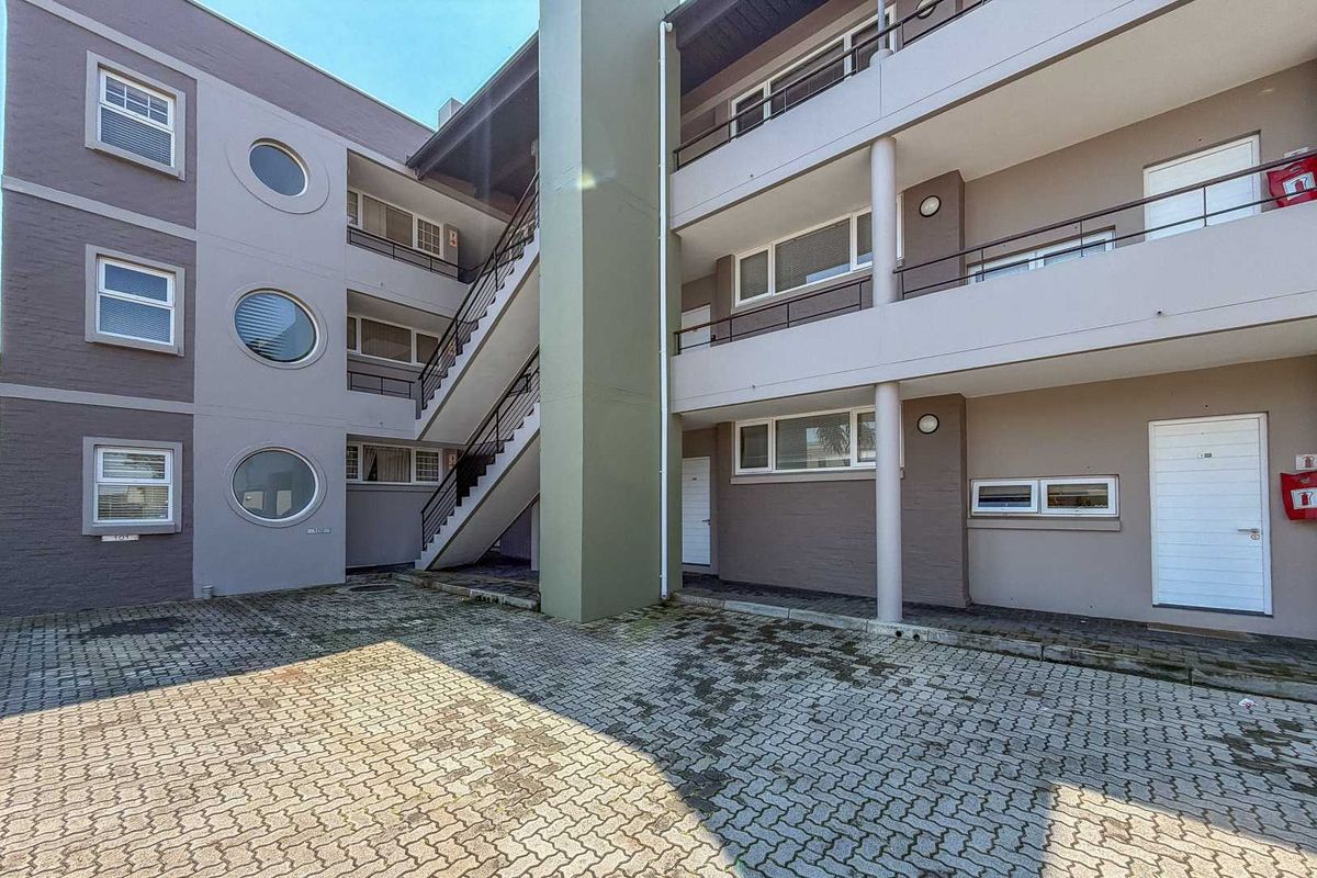 Exterior view of an apartment complex featuring a paved courtyard, external staircase, and multi-level balconies with circular and standard window designs.
