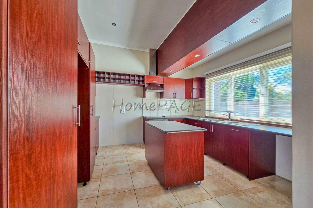 A functional kitchen featuring wood-toned cabinetry, a central island, stainless steel extractor fan, and tiled flooring with ample natural light.