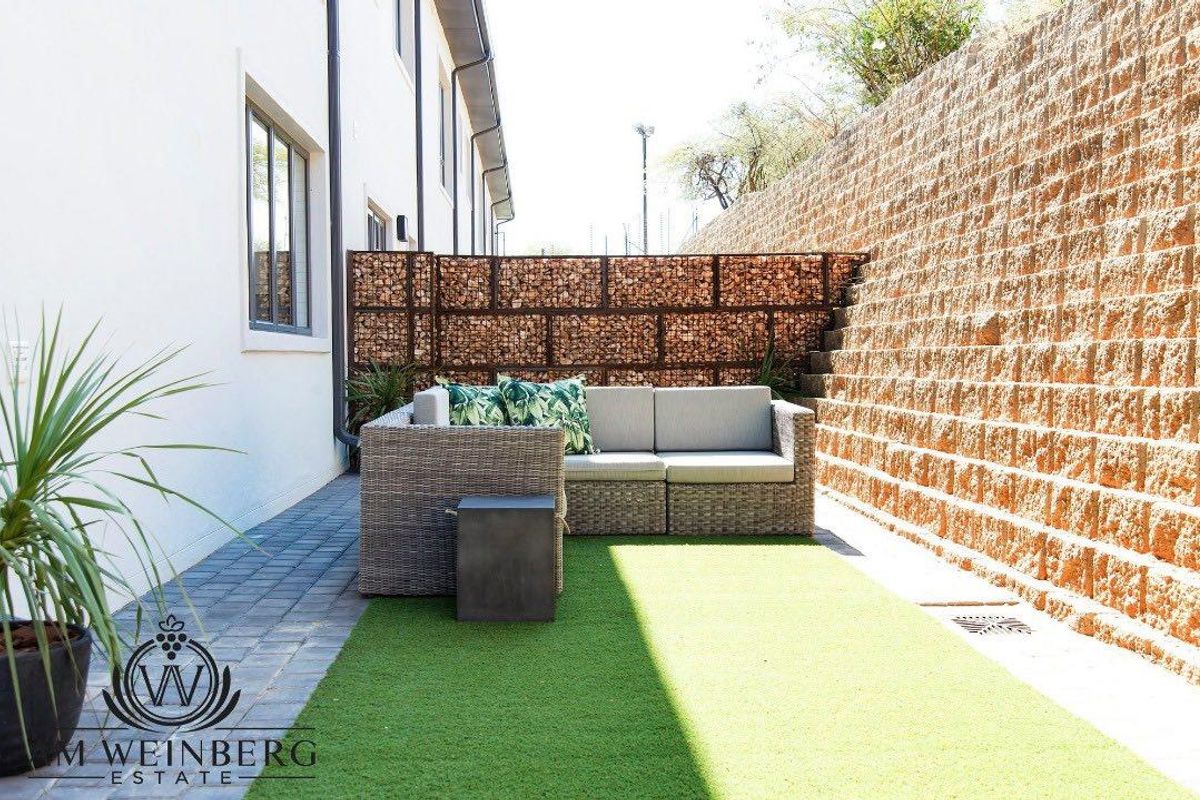 A private rear courtyard featuring a paved walkway, artificial lawn, and a decorative gabion retaining wall.
