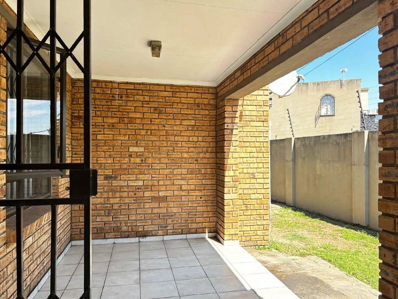 A covered patio area featuring face brick walls, tiled flooring, and a security gate, providing direct access to the outdoor garden space.