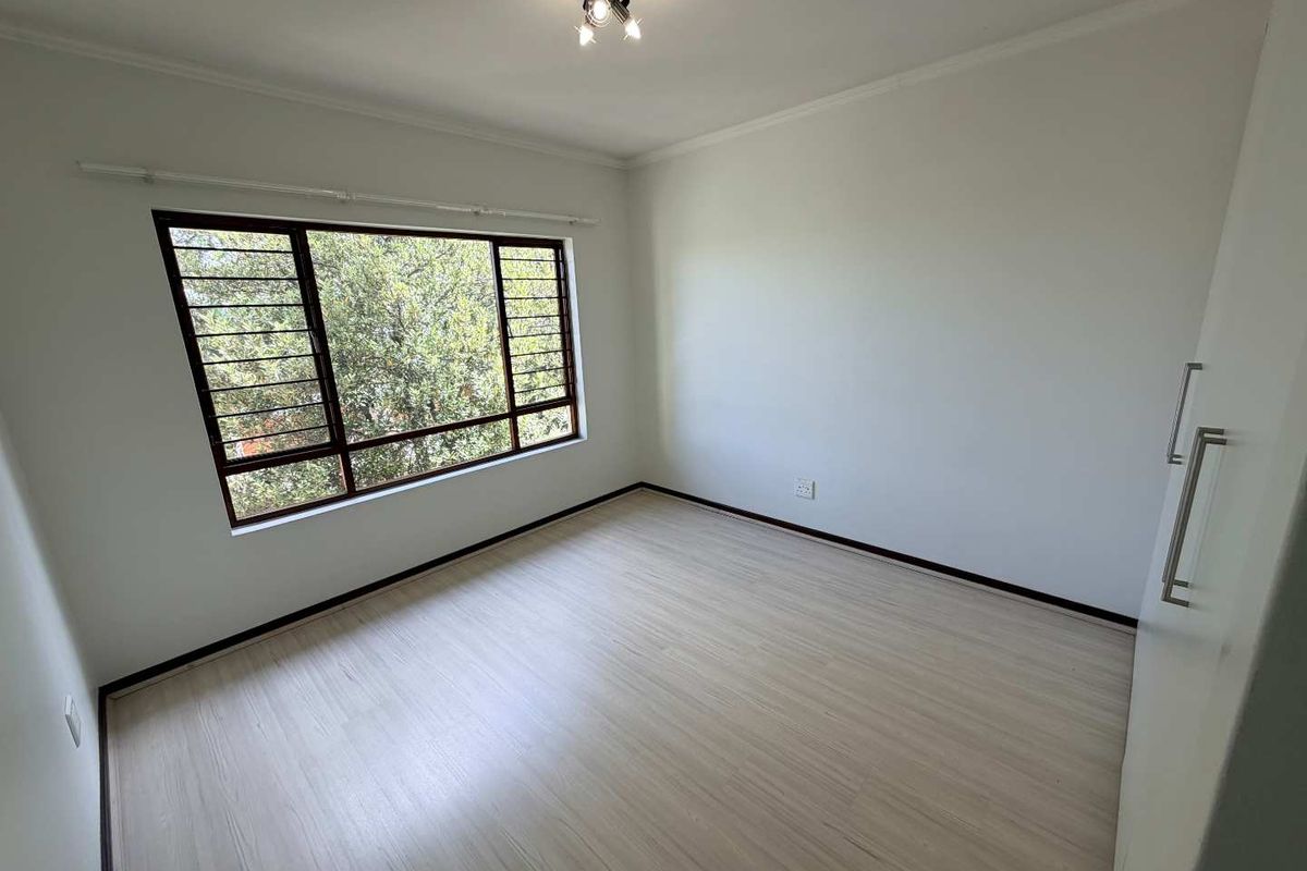 A bright, empty bedroom featuring light-toned laminate flooring, built-in cupboards, and a large window with security bars.