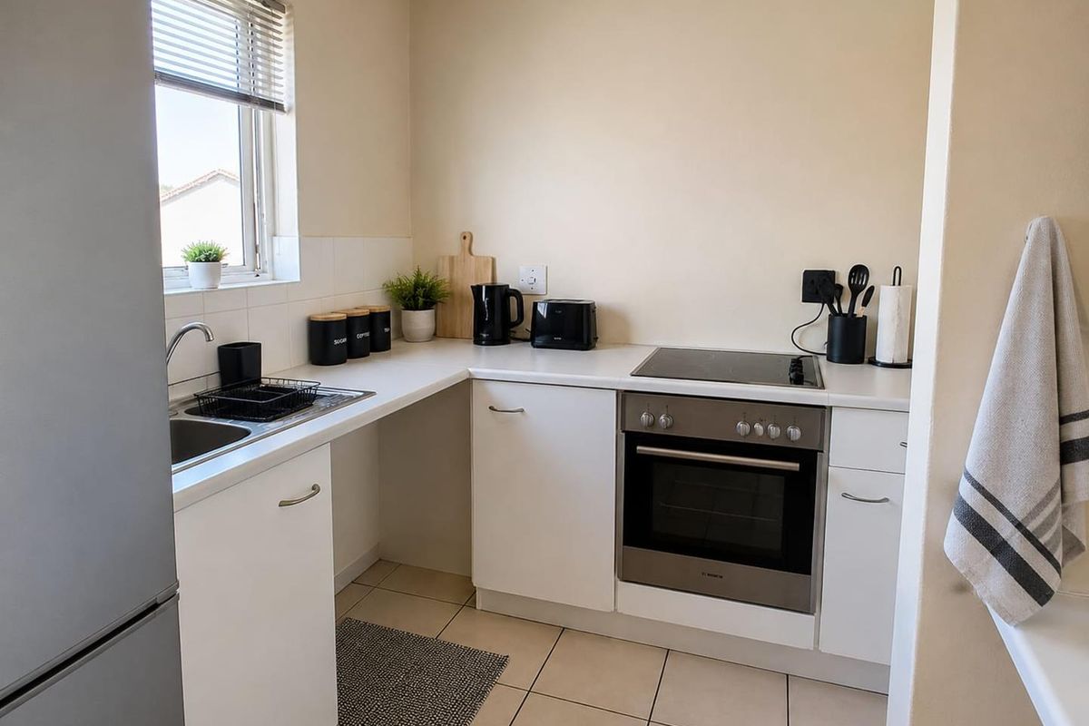 A compact, functional kitchen featuring white cabinetry, a built-in oven with electric hob, stainless steel sink, and neutral tiled flooring.