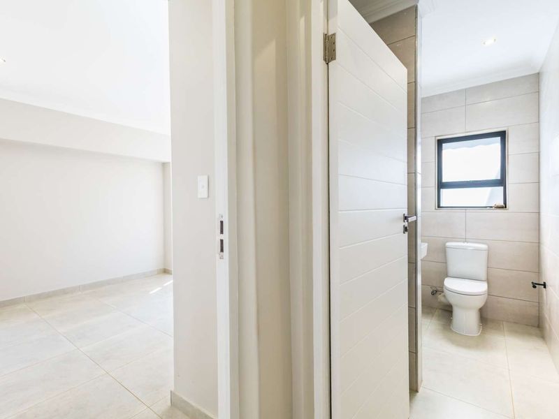 A clean, modern bathroom featuring neutral wall and floor tiles, a standard toilet, and recessed lighting, viewed from an adjacent empty room.
