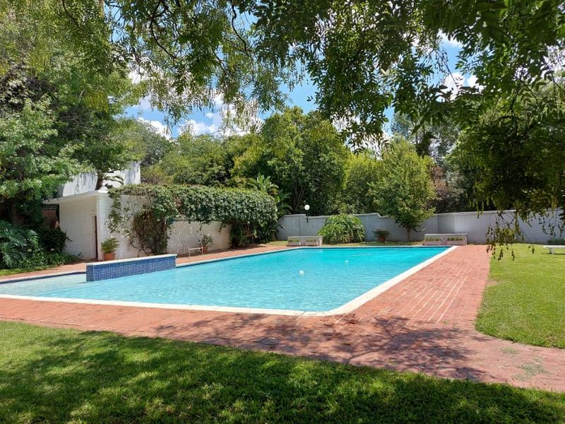 A spacious outdoor swimming pool area featuring a brick-paved surround, landscaped garden, and perimeter walling in a tranquil setting.