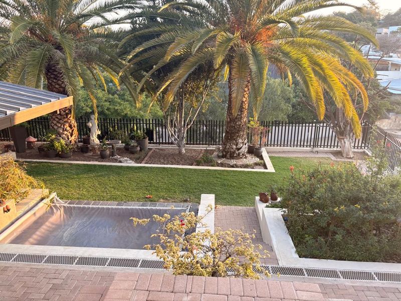 A well-maintained outdoor pool area featuring a covered swimming pool, brick paved surrounds, tiered garden beds, and mature palm trees.