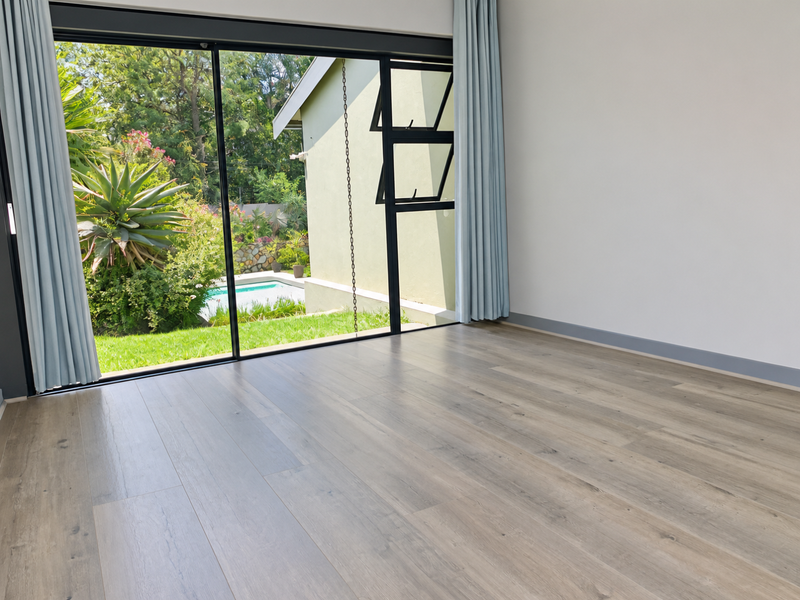 A bright, empty bedroom featuring modern laminate flooring and large sliding glass doors that open to a garden and pool area.