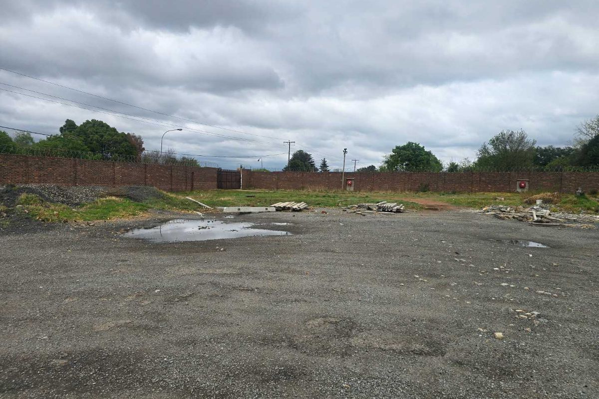 A large, undeveloped gravel yard enclosed by a perimeter brick wall with security razor wire and a metal access gate.