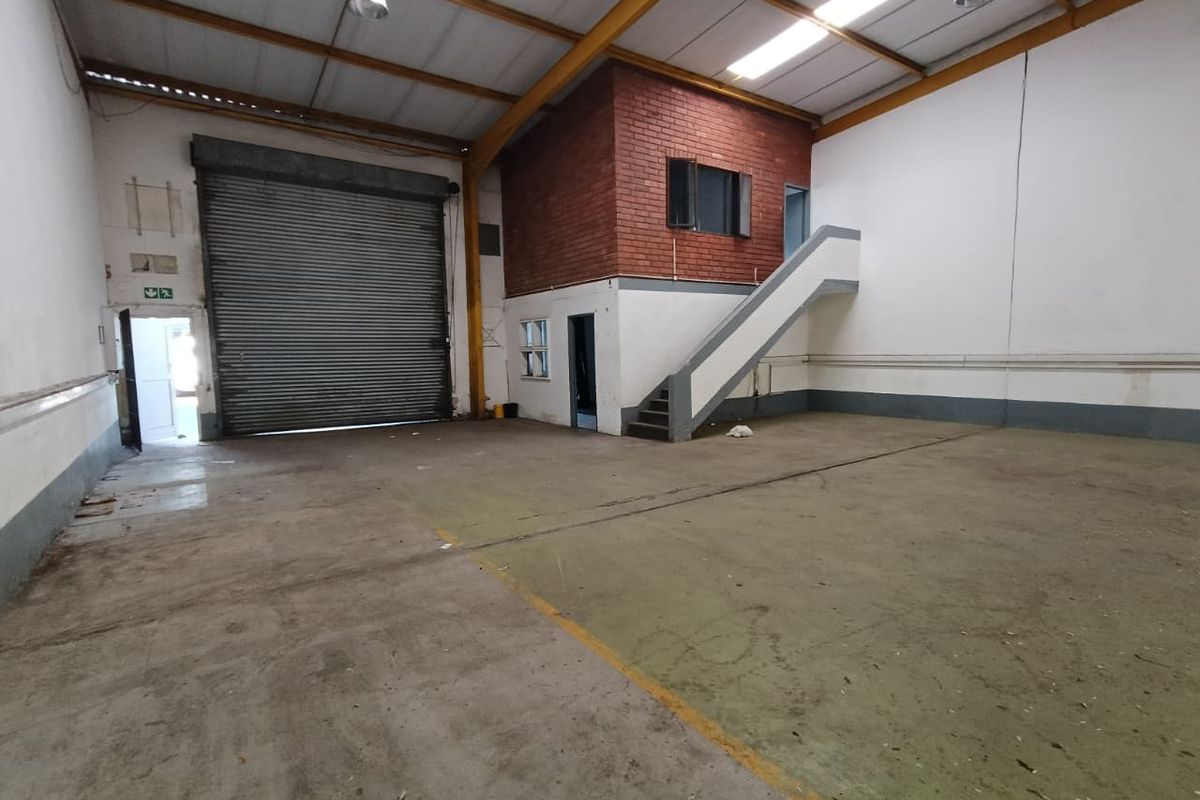 Spacious industrial warehouse unit featuring a high roller shutter door, concrete flooring, and a mezzanine level with an integrated staircase.