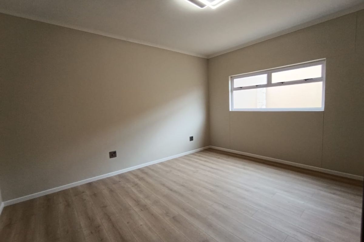 A bright, empty bedroom featuring modern wood-look laminate flooring, neutral wall finishes, and a contemporary square LED ceiling light fixture.