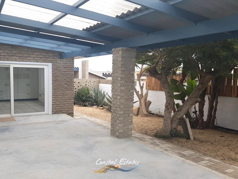 A covered outdoor patio area featuring brick columns, translucent roofing, and sliding door access to the interior, overlooking a landscaped garden space.