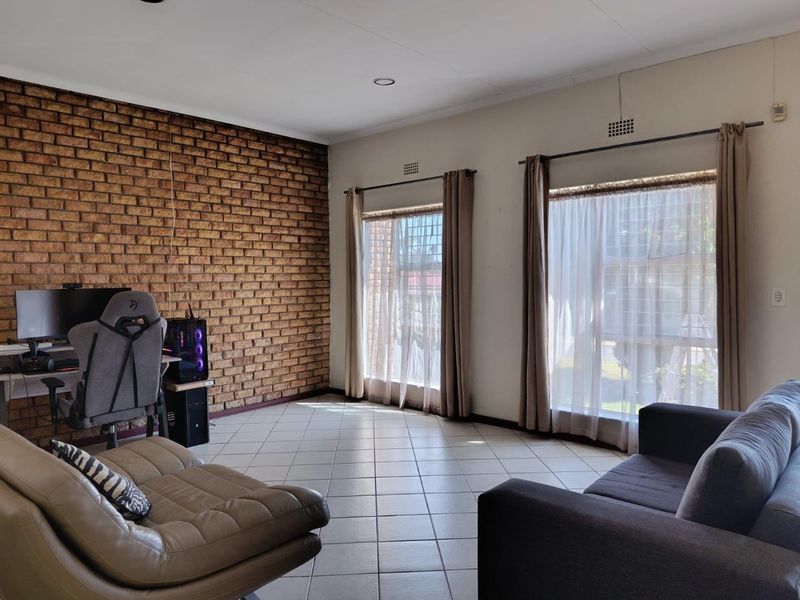 Spacious living room featuring a prominent exposed brick wall, tiled flooring, and large windows with natural light.