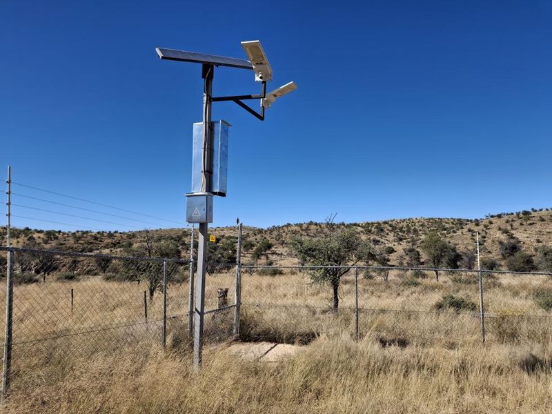 A security installation featuring solar-powered lighting and cameras mounted on a pole, situated within a fenced perimeter on the small holding.