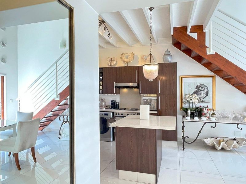 Open plan kitchen and living area featuring dark wood cabinetry, stone island, white tiled floors, and a striking timber staircase with white railings.