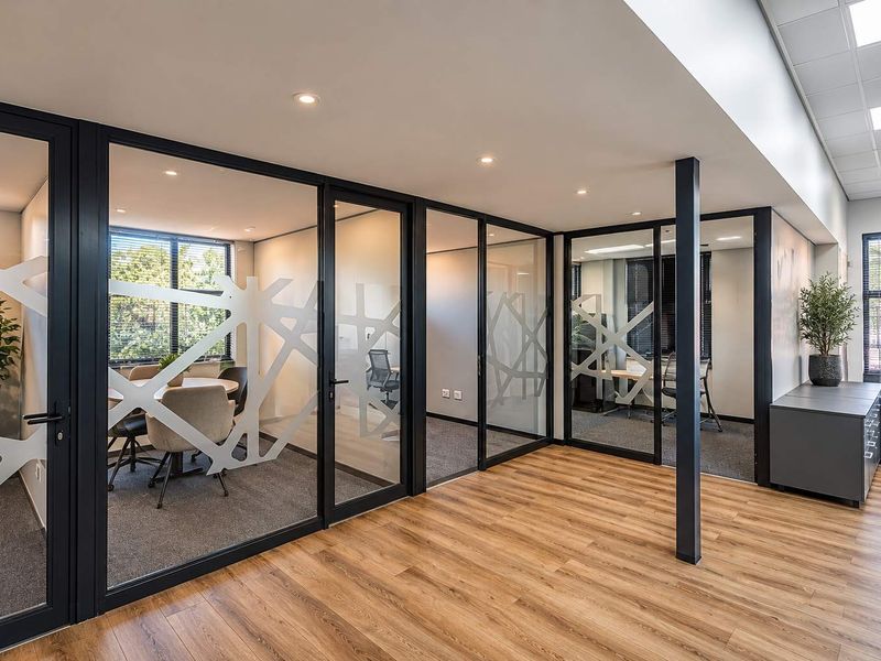 Modern office space featuring glass-partitioned meeting rooms, black aluminum framing, recessed lighting, and a combination of wood-look vinyl and carpeted flooring.