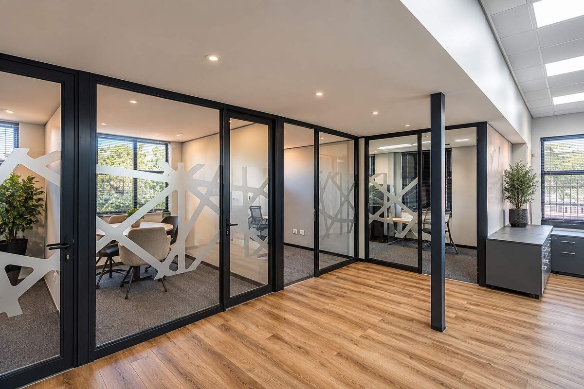 Modern office space featuring glass-partitioned meeting rooms, black aluminum framing, recessed lighting, and a combination of wood-look vinyl and carpeted flooring.