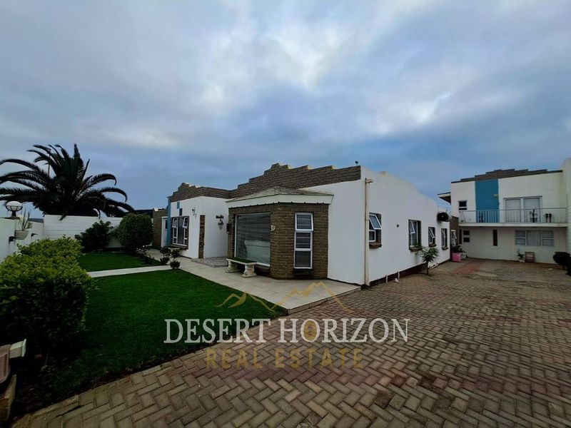 Front view of a residential property featuring a paved driveway, landscaped garden, and secure exterior finishes with security shutters.