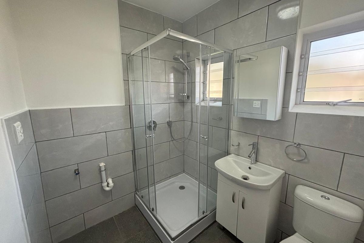 A clean, modern bathroom featuring a glass shower enclosure, vanity basin, and toilet with neutral grey wall tiling and louver windows.