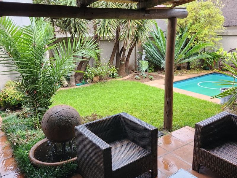 Covered patio area featuring a rustic thatched roof and timber beams, overlooking a private garden with a swimming pool and established landscaping.