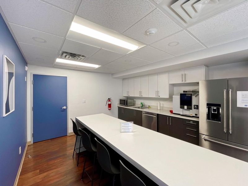 A modern office kitchenette featuring white and dark cabinetry, stainless steel appliances, a central island, and professional drop ceiling lighting.