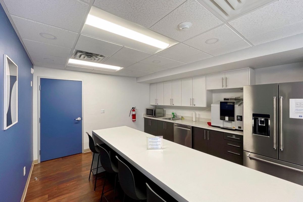 A modern office kitchenette featuring white and dark cabinetry, stainless steel appliances, a central island, and professional drop ceiling lighting.