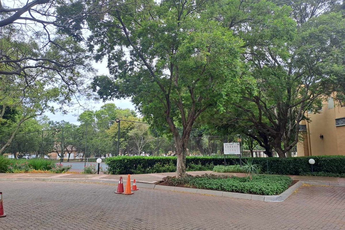 A view of the apartment complex grounds featuring paved driveway, mature trees, landscaped garden beds, and a fenced sports facility in the background.