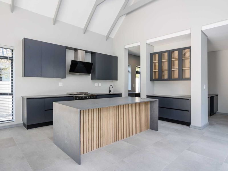 Modern kitchen featuring a central island with timber slat detailing, dark cabinetry, high vaulted ceilings, and sleek tiled flooring in a contemporary open-plan layout.